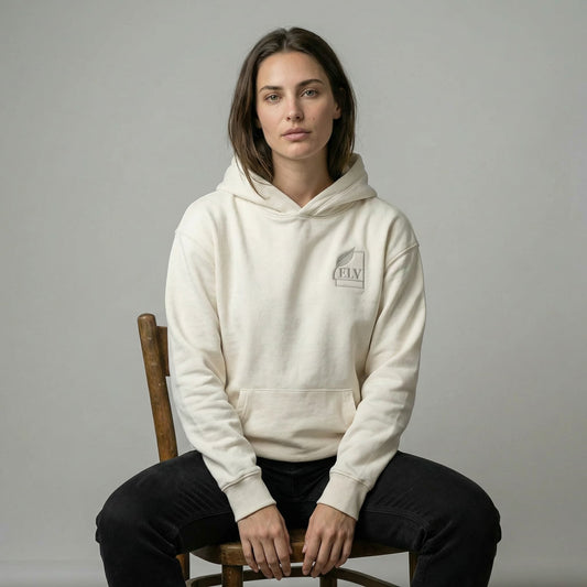 Person wearing a beige hoodie with a logo, sitting on a wooden chair against a plain background